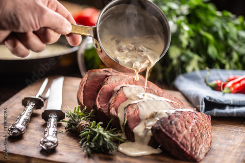 Rump steak cut to slices on a chopping board next to a knife and rosemary, with a mushroom sauce poured over it