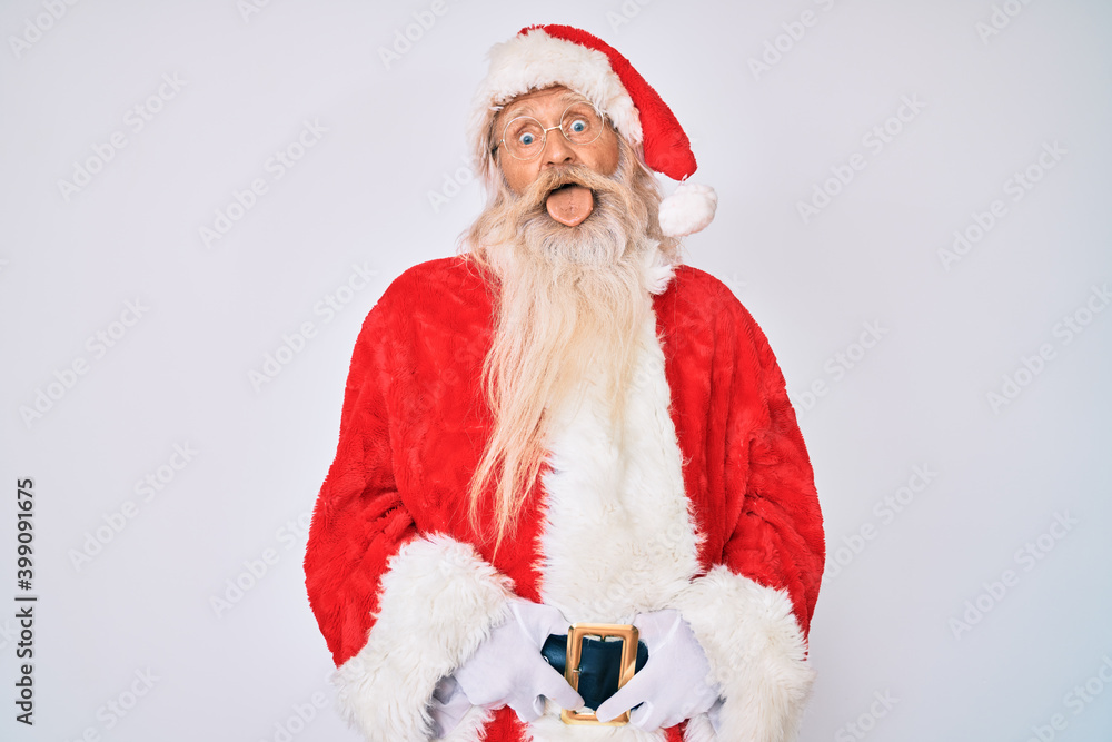 Old senior man with grey hair and long beard wearing santa claus costume with suspenders sticking tongue out happy with funny expression. emotion concept.