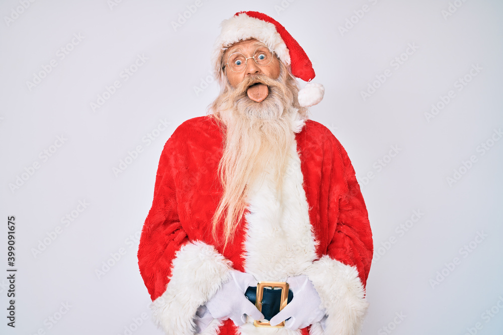 Old senior man with grey hair and long beard wearing santa claus costume with suspenders sticking tongue out happy with funny expression. emotion concept. Old senior man with grey hair and long beard wearing santa claus costume with suspenders sticking tongue out happy with funny expression. emotion concept.