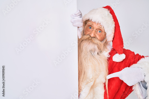 Old senior man with grey hair and long beard wearing santa claus costume holding banner pointing finger to one self smiling happy and proud