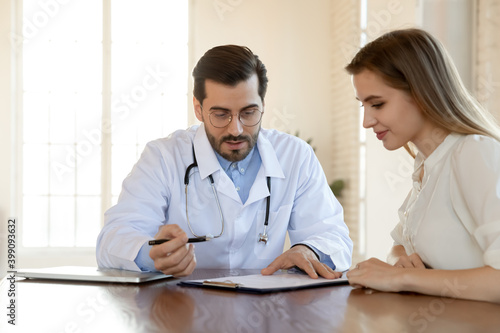 Young male doctor in white medical uniform sit at desk in clinic with female patient discuss illness anamnesis together. Man GP or physician talk consult woman client, consider results or treatment.