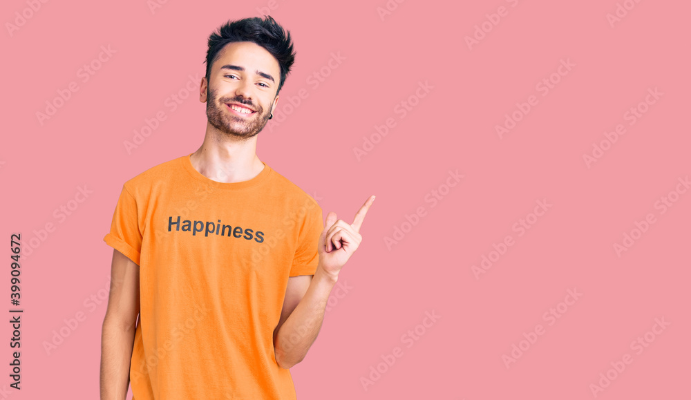 Young hispanic man wearing t shirt with happiness word message cheerful with a smile on face pointing with hand and finger up to the side with happy and natural expression Young hispanic man wearing t shirt with happiness word message cheerful with a smile on face pointing with hand and finger up to the side with happy and natural expression