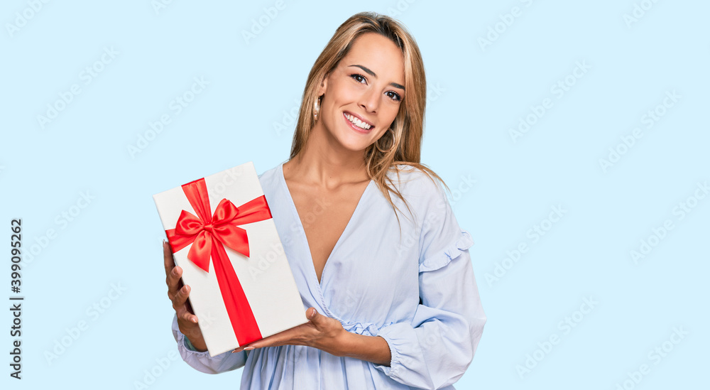 Beautiful blonde woman holding gift looking positive and happy standing and smiling with a confident smile showing teeth Beautiful blonde woman holding gift looking positive and happy standing and smiling with a confident smile showing teeth