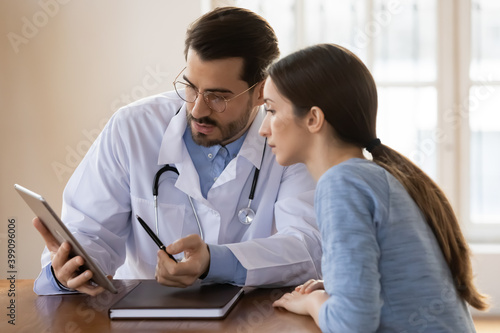 Young Caucasian male doctor and female patient look at tablet screen discuss test results on gadget together. Man GP or physician use pad explain anamnesis to woman client at consultation in hospital.