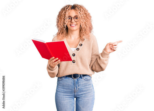 Young blonde woman with curly hair wearing glasses and reading book smiling happy pointing with hand and finger to the side