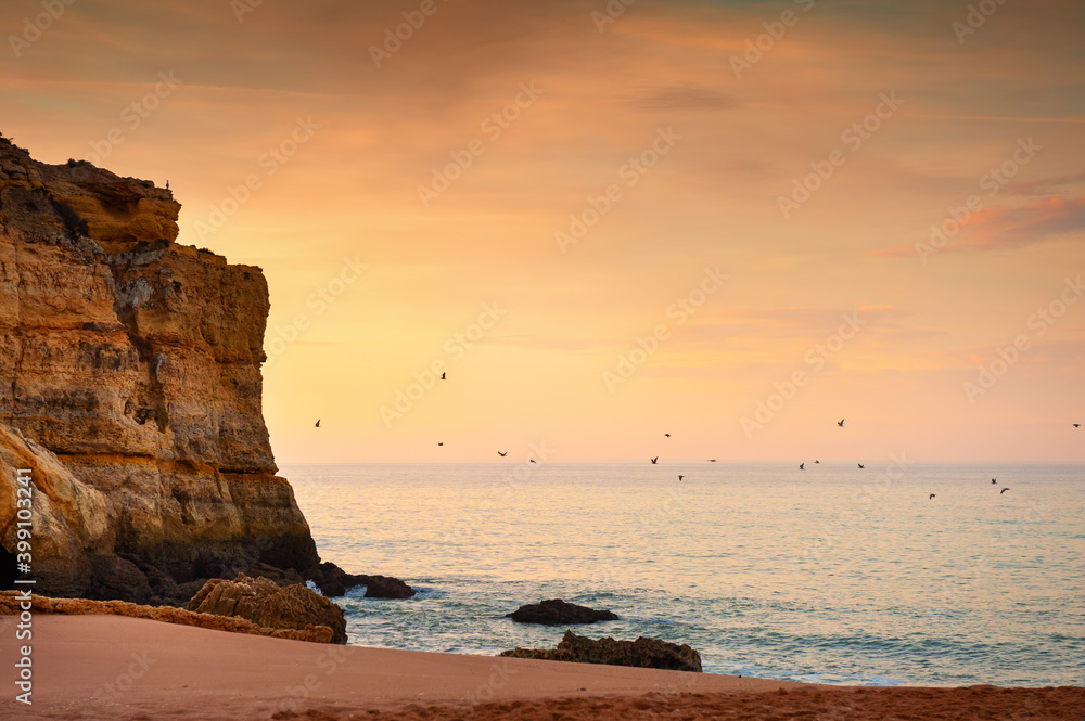 Beautiful sunrise on the beach in Algarve, Portugal. Flock of birds flying over the water. Shore of Atlantic ocean. Beautiful summer seascape, famous travel destination