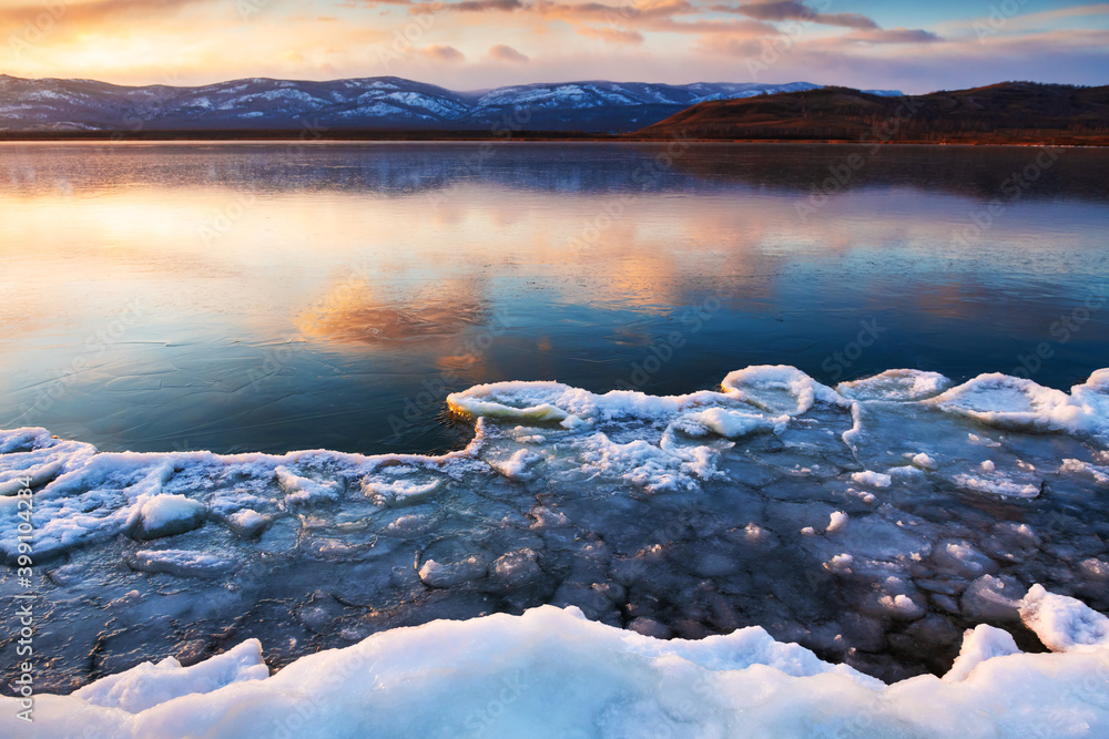 Ice on the frozen lake at sunset. Mountains and sky are reflected in the ice surface of the lake. Beautiful winter landscape. South Ural, Russia - obrazy, fototapety, plakaty 