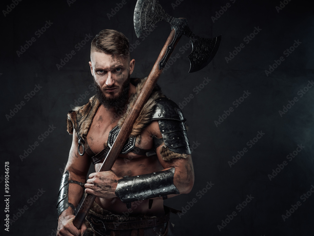 Dressed in dark armour with fur dirty and awesome viking fighter with two handed axe on his shoulder poses in dark background.