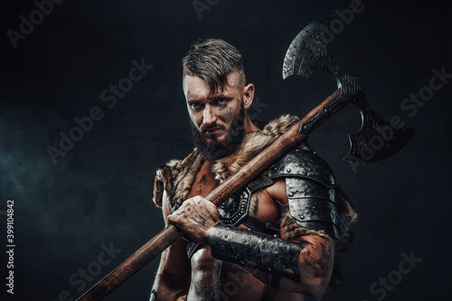 Holding two handed axe on his shoulder scandinavian barbarian in light armour with fur poses in dark background looking at camera. photo