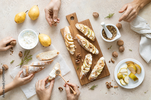 Woman, man and child are cooking sweet open sandwiches with ricotta cheese, fresh pears, walnuts and honey on kitchen table for family breakfast, overhead view