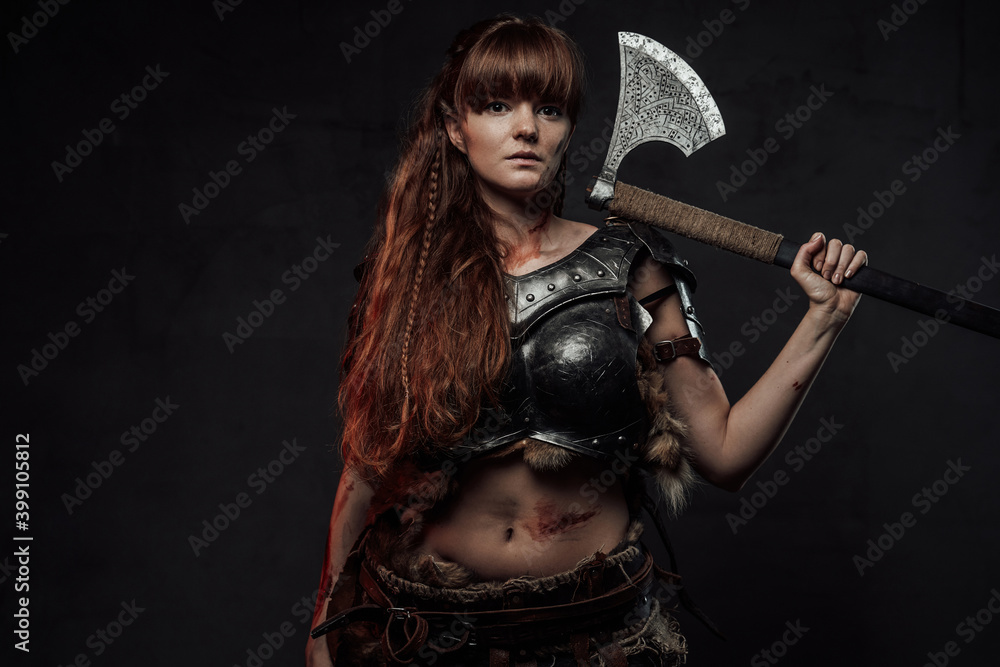Brown haired beautiful northern amazon posing in dark background with hatchet on her shoulder.