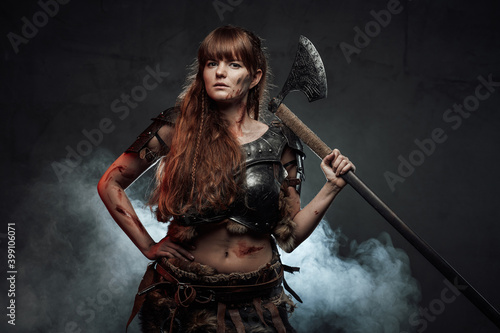 Barbaric and brown haired woman viking wielding two handed axe and dressed in dark armour poses in dark smokey background.