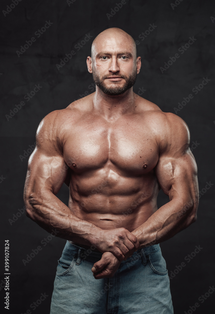 Caucasian topless guy with bald head and muscular build in jeans shorts poses in dark background looking at camera.