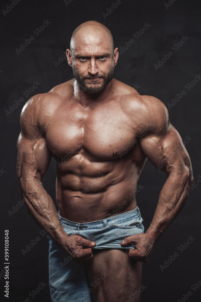 Caucasian muscular man with hairless head and naked torso pulls his shorts showing his muscular leg looking at camera with serious face in dark background.
