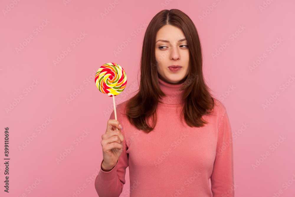 Happy brunette woman in pink sweater looking with desire at striped colorful lollipop on stick in her hand, crazy about sweet confectionery. Indoor studio shot isolated on pink background