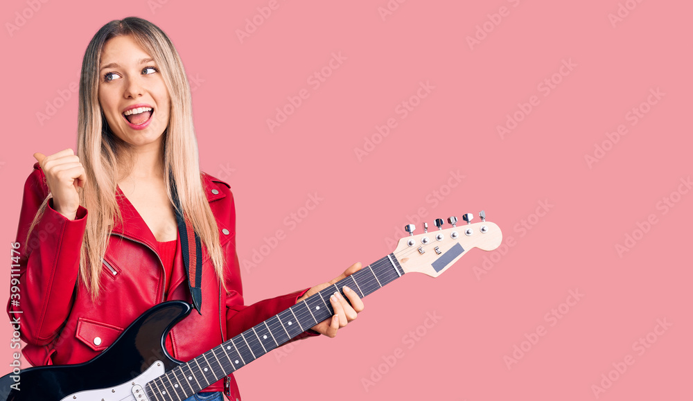 Young beautiful blonde woman playing electric guitar pointing thumb up to the side smiling happy with open mouth Young beautiful blonde woman playing electric guitar pointing thumb up to the side smiling happy with open mouth