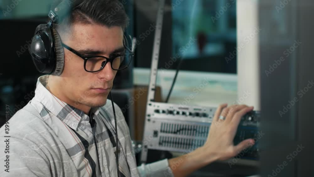 A young man with glasses and headphones adjusts the volume on the control panel in a recording studio. The sound engineer of the TV channel controls the sound volume while sitting at the workplace. A young man with glasses and headphones adjusts the volume on the control panel in a recording studio. The sound engineer of the TV channel controls the sound volume while sitting at the workplace.