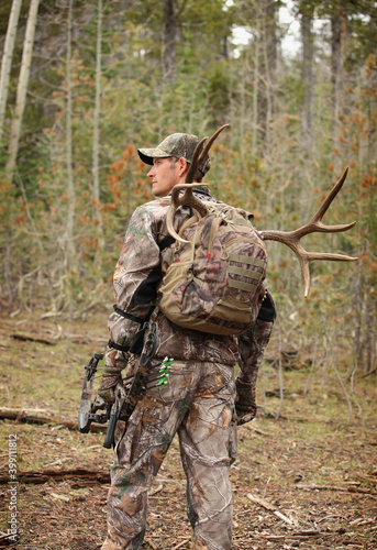 successful bow hunter hiking through woods with deer in backpack
