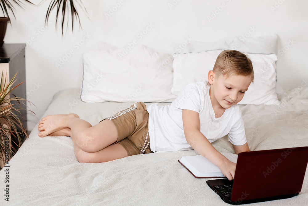 blonde eight-year-old boy studies at home online with a laptop. The child on the bed does homework remotely. blonde eight-year-old boy studies at home online with a laptop. The child on the bed does homework remotely.
