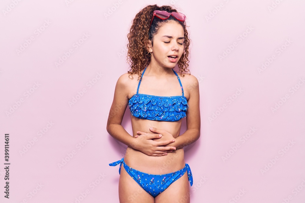 Naklejka premium Beautiful kid girl with curly hair wearing bikini and sunglasses with hand on stomach because nausea, painful disease feeling unwell. ache concept.