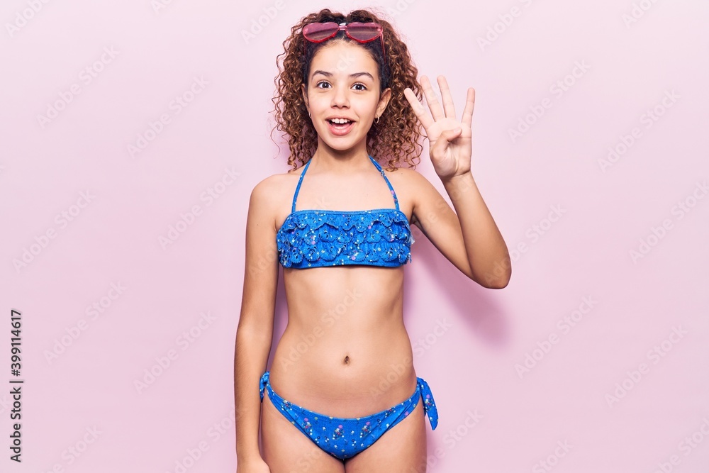 Naklejka premium Beautiful kid girl with curly hair wearing bikini and sunglasses showing and pointing up with fingers number four while smiling confident and happy.