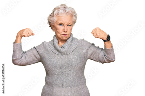 Senior grey-haired woman wearing casual winter sweater showing arms muscles smiling proud. fitness concept.