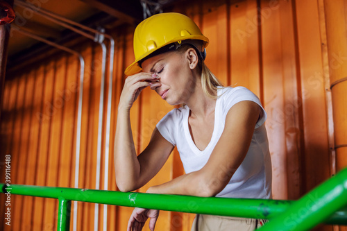 Exhausted female worker with protective helmet on head leaning on railing in factory and having headache because she is overworked. photo