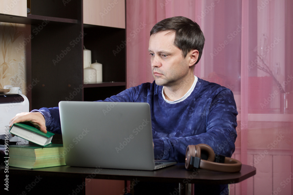 A man in home clothes works at a laptop. Books and headphones are on the table nearby. A man in home clothes works at a laptop. Books and headphones are on the table nearby.