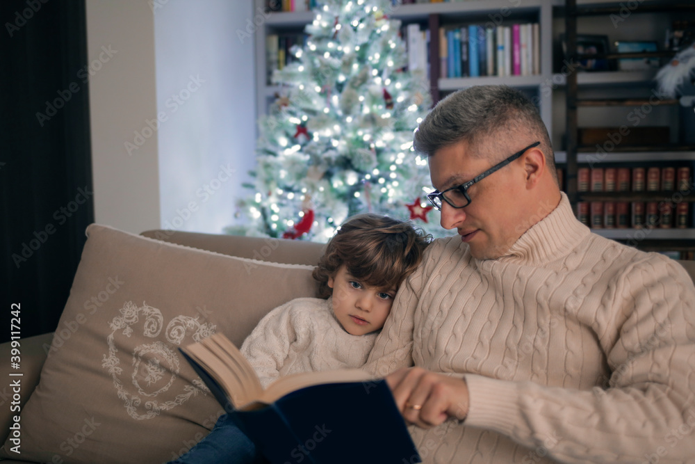 Dad and son are reading a book near the Christmas three Dad and son are reading a book near the Christmas three