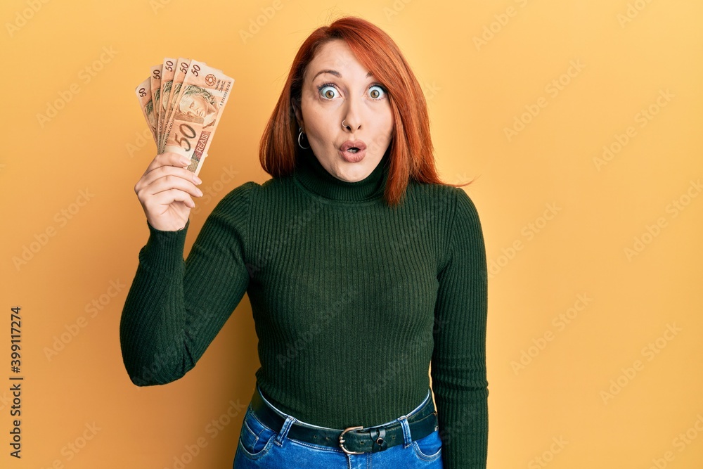 Beautiful redhead woman holding 50 brazilian real banknotes scared and amazed with open mouth for surprise, disbelief face Beautiful redhead woman holding 50 brazilian real banknotes scared and amazed with open mouth for surprise, disbelief face