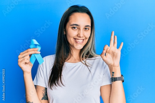 Young hispanic woman holding blue ribbon doing ok sign with fingers  smiling friendly gesturing excellent symbol