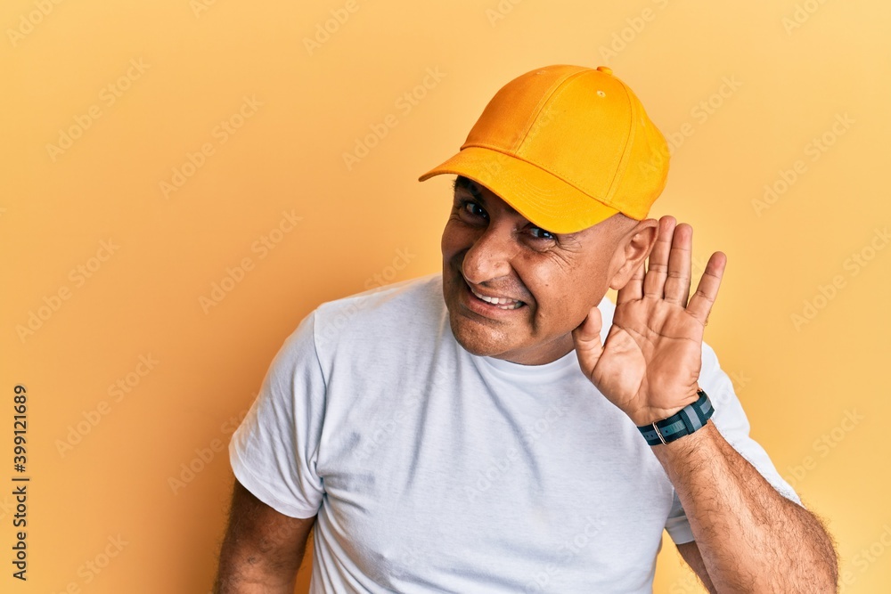 Mature middle east man with mustache wearing casual white tshirt and yellow cap smiling with hand over ear listening and hearing to rumor or gossip. deafness concept. Mature middle east man with mustache wearing casual white tshirt and yellow cap smiling with hand over ear listening and hearing to rumor or gossip. deafness concept.