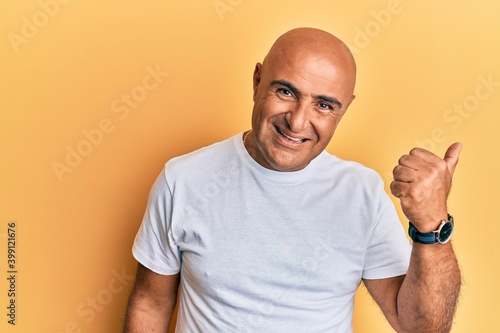 Mature middle east man wearing casual white tshirt smiling with happy face looking and pointing to the side with thumb up.