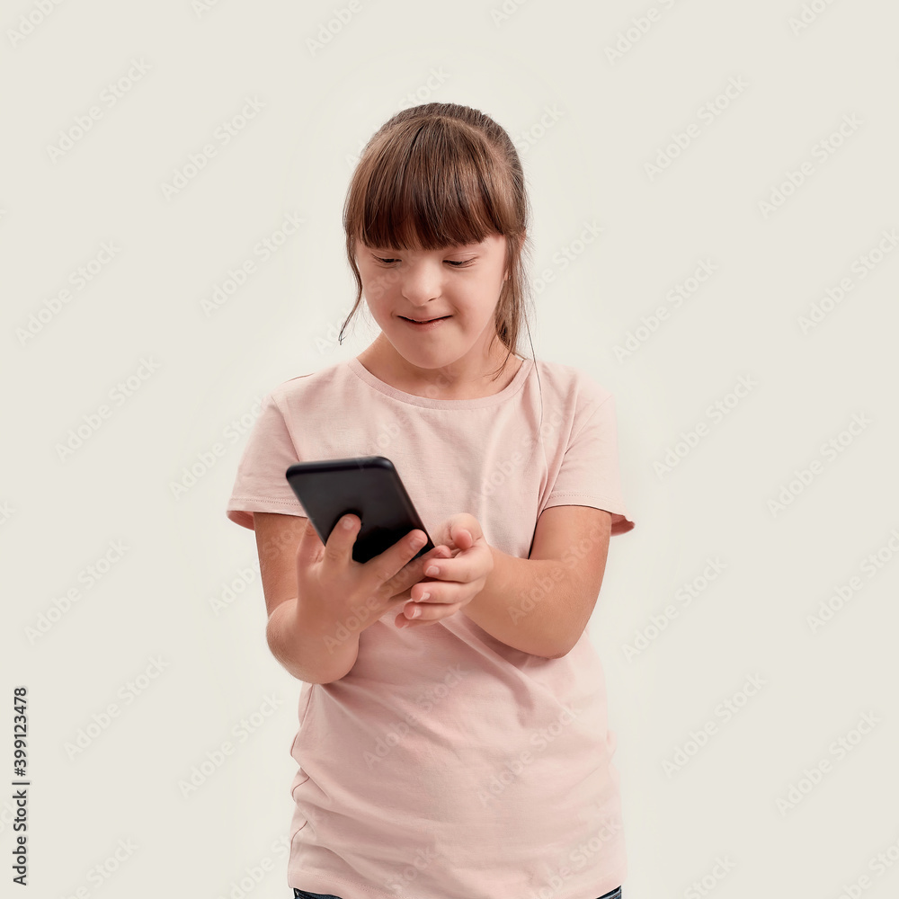 Portrait of disabled girl with Down syndrome holding smartphone, looking at the screen while posing isolated over white background Portrait of disabled girl with Down syndrome holding smartphone, looking at the screen while posing isolated over white background