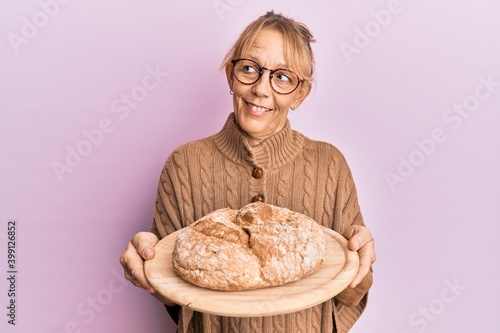 Middle age blonde woman holding bread smiling looking to the side and staring away thinking.