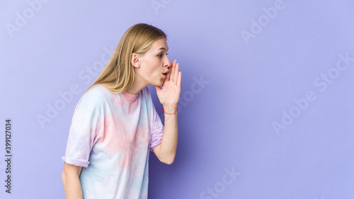 Young blonde woman isolated on purple background is saying a secret hot braking news and looking aside