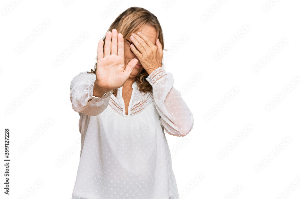 Middle age blonde woman wearing casual white shirt and glasses covering eyes with hands and doing stop gesture with sad and fear expression. embarrassed and negative concept. Middle age blonde woman wearing casual white shirt and glasses covering eyes with hands and doing stop gesture with sad and fear expression. embarrassed and negative concept.