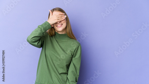 Young blonde woman isolated on purple background covers eyes with hands, smiles broadly waiting for a surprise.