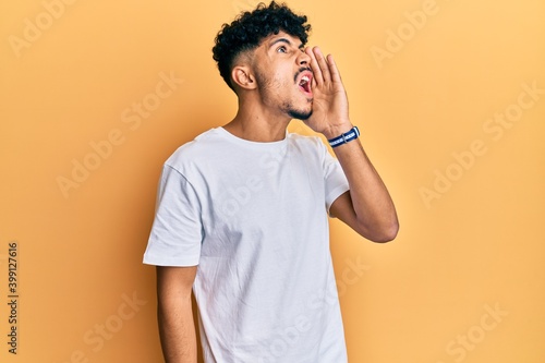 Young arab handsome man wearing casual white tshirt shouting and screaming loud to side with hand on mouth. communication concept.