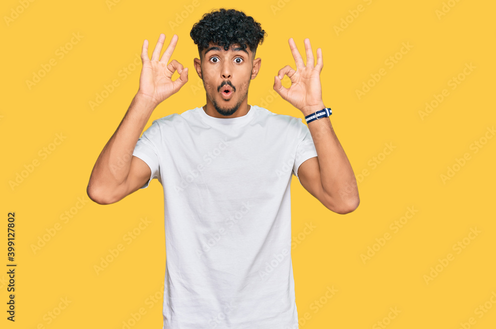 Young arab man wearing casual white t shirt looking surprised and shocked doing ok approval symbol with fingers. crazy expression Young arab man wearing casual white t shirt looking surprised and shocked doing ok approval symbol with fingers. crazy expression