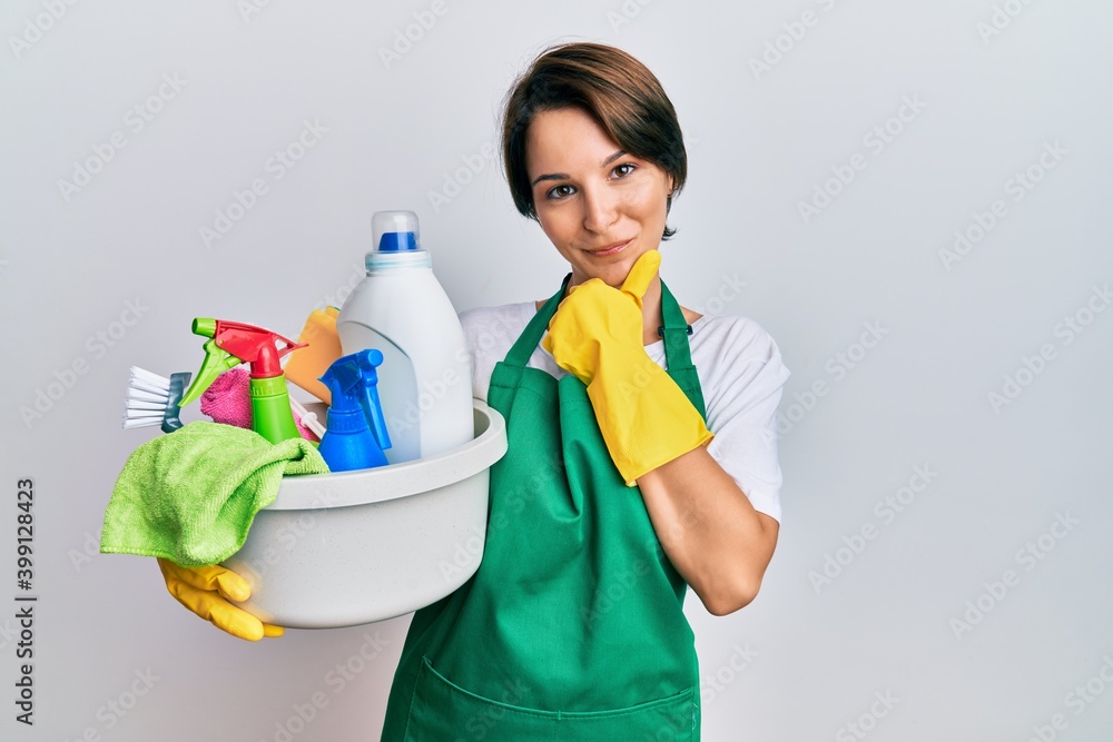 Young brunette woman with short hair wearing apron holding cleaning products looking confident at the camera smiling with crossed arms and hand raised on chin. thinking positive. Young brunette woman with short hair wearing apron holding cleaning products looking confident at the camera smiling with crossed arms and hand raised on chin. thinking positive.