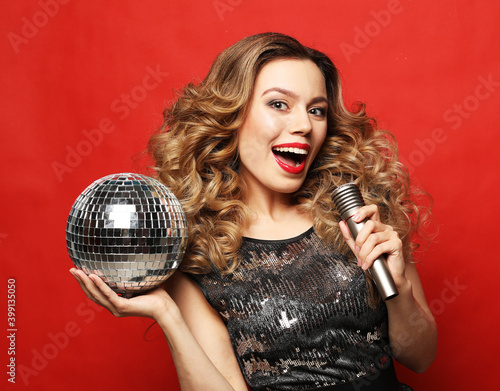 Young blond woman with long wavy hair dressed in evening dress holding a microphone and disco ball  singing and smiling