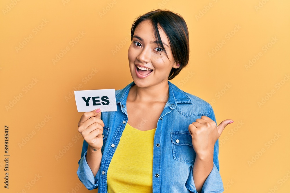 Beautiful young woman with short hair holding yes reminder pointing thumb up to the side smiling happy with open mouth Beautiful young woman with short hair holding yes reminder pointing thumb up to the side smiling happy with open mouth