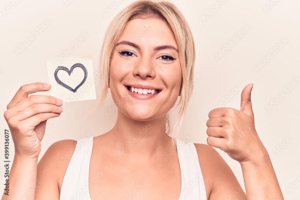 Young beautiful blonde woman holding reminder paper with heart shape over white background smiling happy and positive, thumb up doing excellent and approval sign Young beautiful blonde woman holding reminder paper with heart shape over white background smiling happy and positive, thumb up doing excellent and approval sign