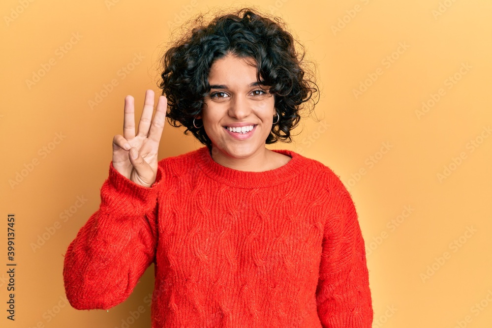 Young hispanic woman with curly hair wearing casual winter sweater showing and pointing up with fingers number three while smiling confident and happy. Young hispanic woman with curly hair wearing casual winter sweater showing and pointing up with fingers number three while smiling confident and happy.