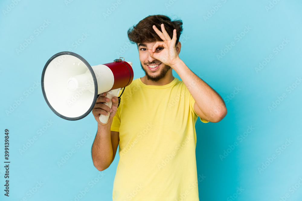 Young arab man holding a megaphone excited keeping ok gesture on eye. Young arab man holding a megaphone excited keeping ok gesture on eye.