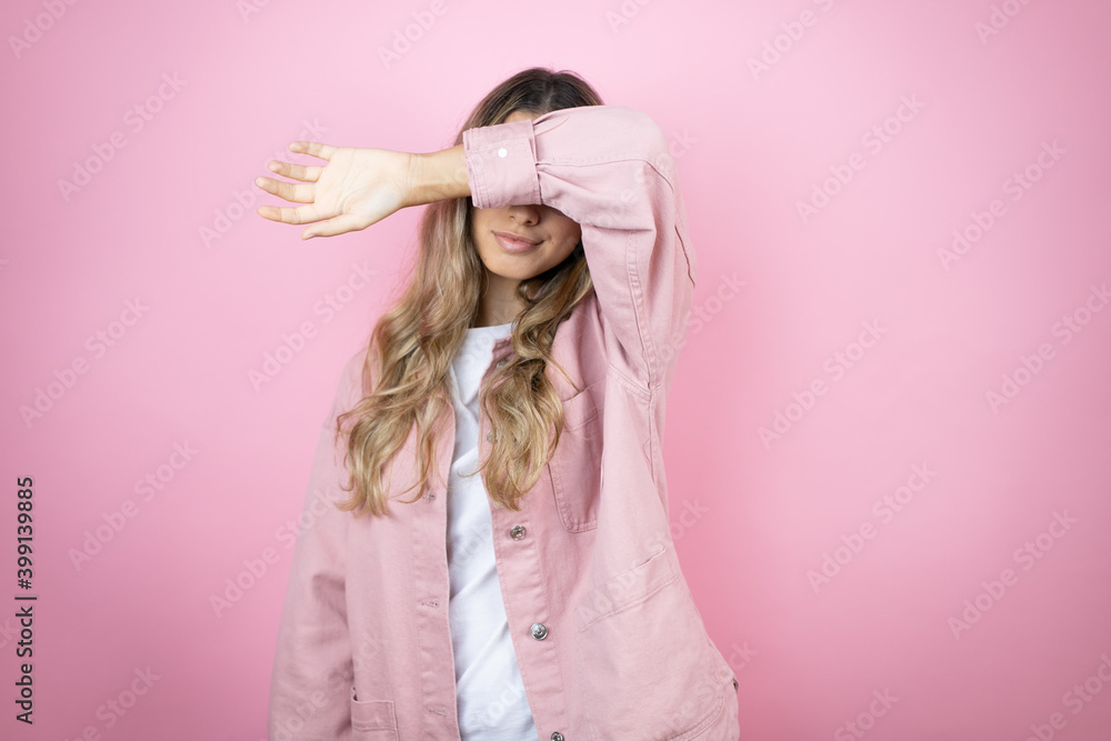 Young beautiful blonde woman with long hair standing over pink background covering eyes with arm smiling cheerful and funny. Blind concept. Young beautiful blonde woman with long hair standing over pink background covering eyes with arm smiling cheerful and funny. Blind concept.