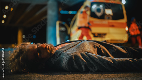 Shot of a Young of a Bloody Man Lying on the Pavement after Traffic Accident on a Street at Night. Team of EMS Paramedics Quickly Jump Out from Ambulance Vehicle. Emergency Care Assistants Run.