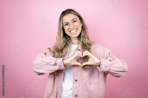 Young beautiful blonde woman with long hair standing over pink background smiling in love showing heart symbol and shape with hands