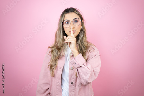 Young beautiful blonde woman with long hair standing over pink background asking to be quiet with finger on lips. Silence and secret concept.